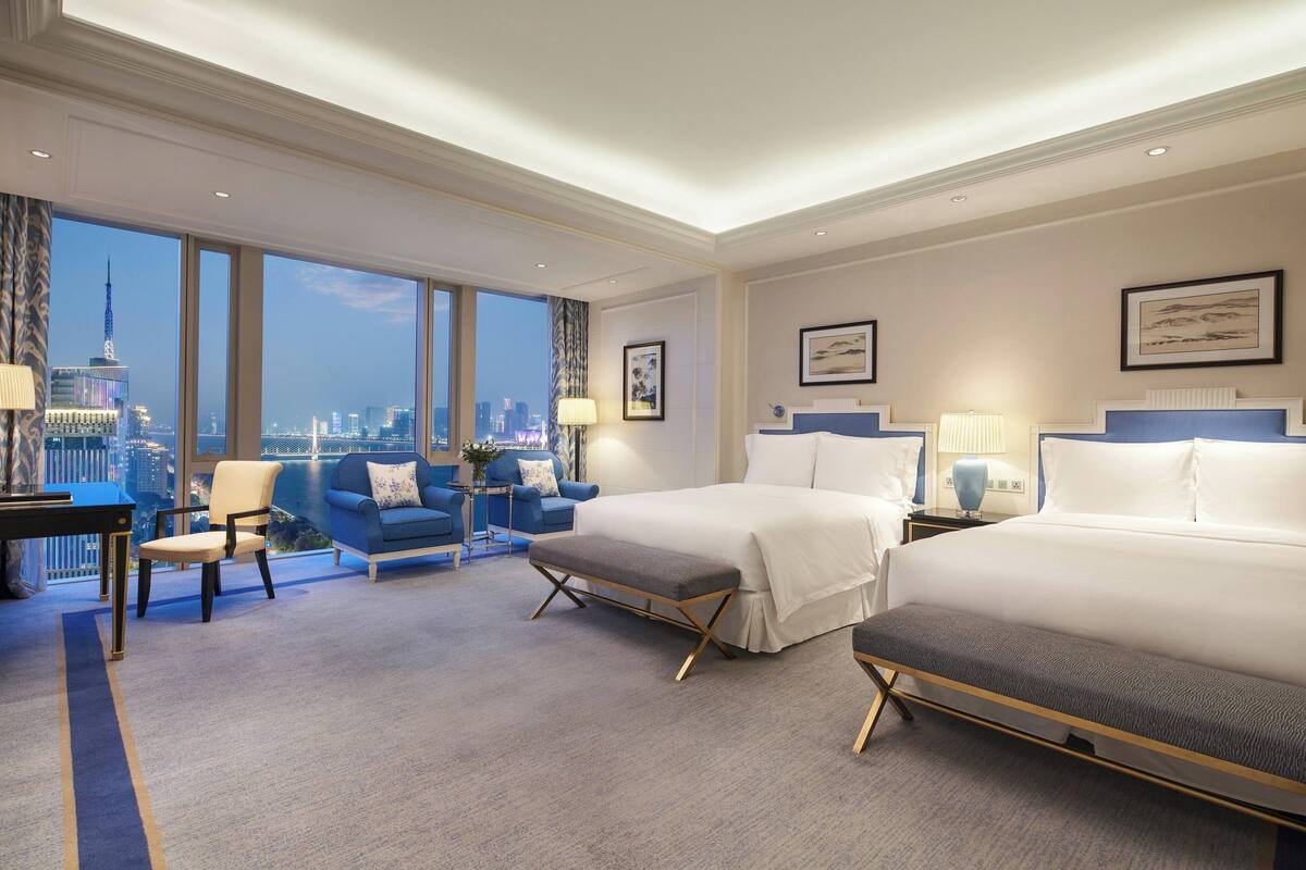 A hotel room with two beds, a desk, a chair, and a view of the cityscape.