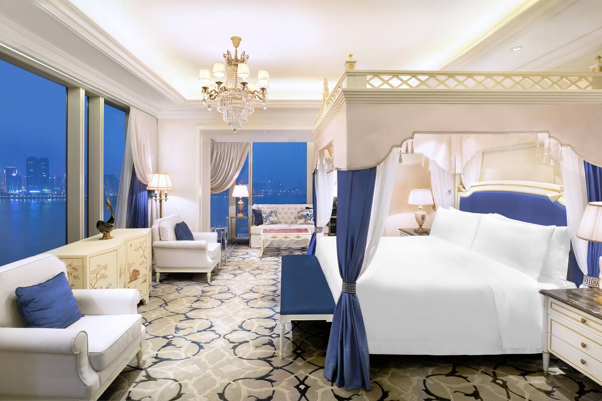 A luxurious bedroom with a large bed, a sitting area with a sofa and armchairs, a nightstand, and a view of the cityscape.