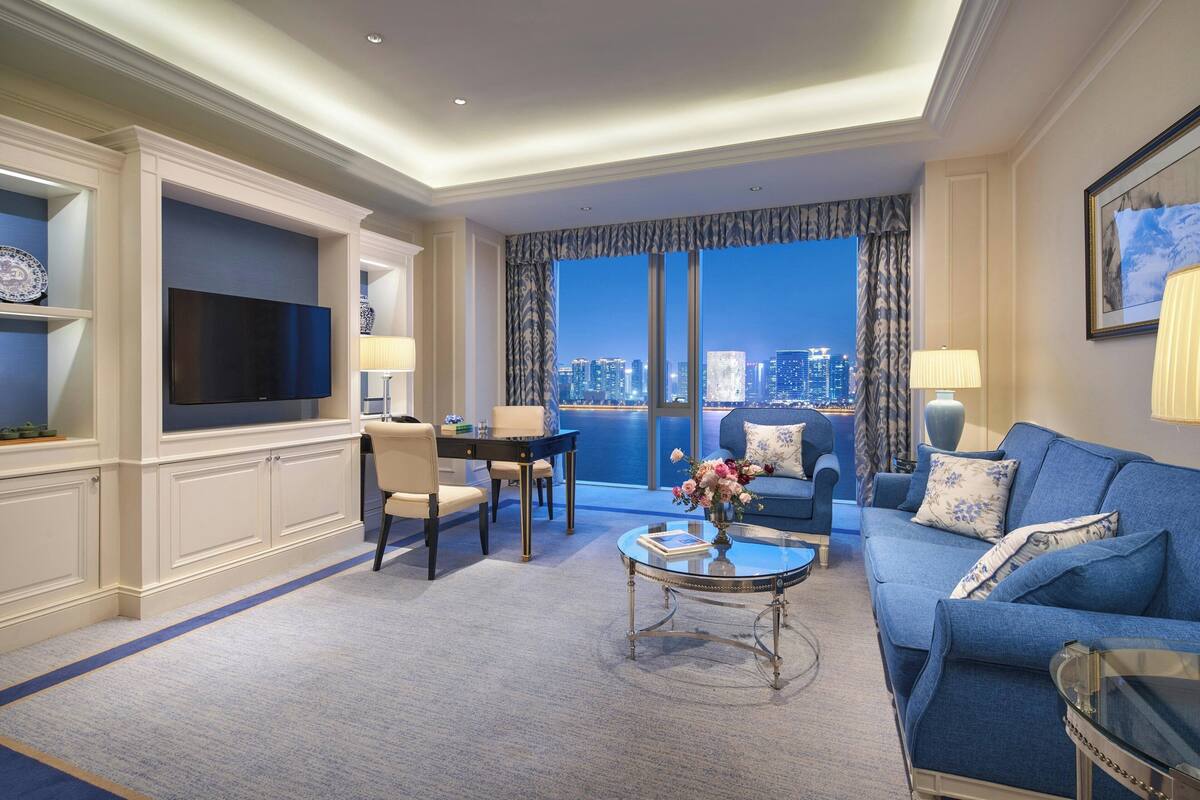 A modern hotel room with a large flat-screen TV, a sofa, a dining area, and a view of the city skyline.