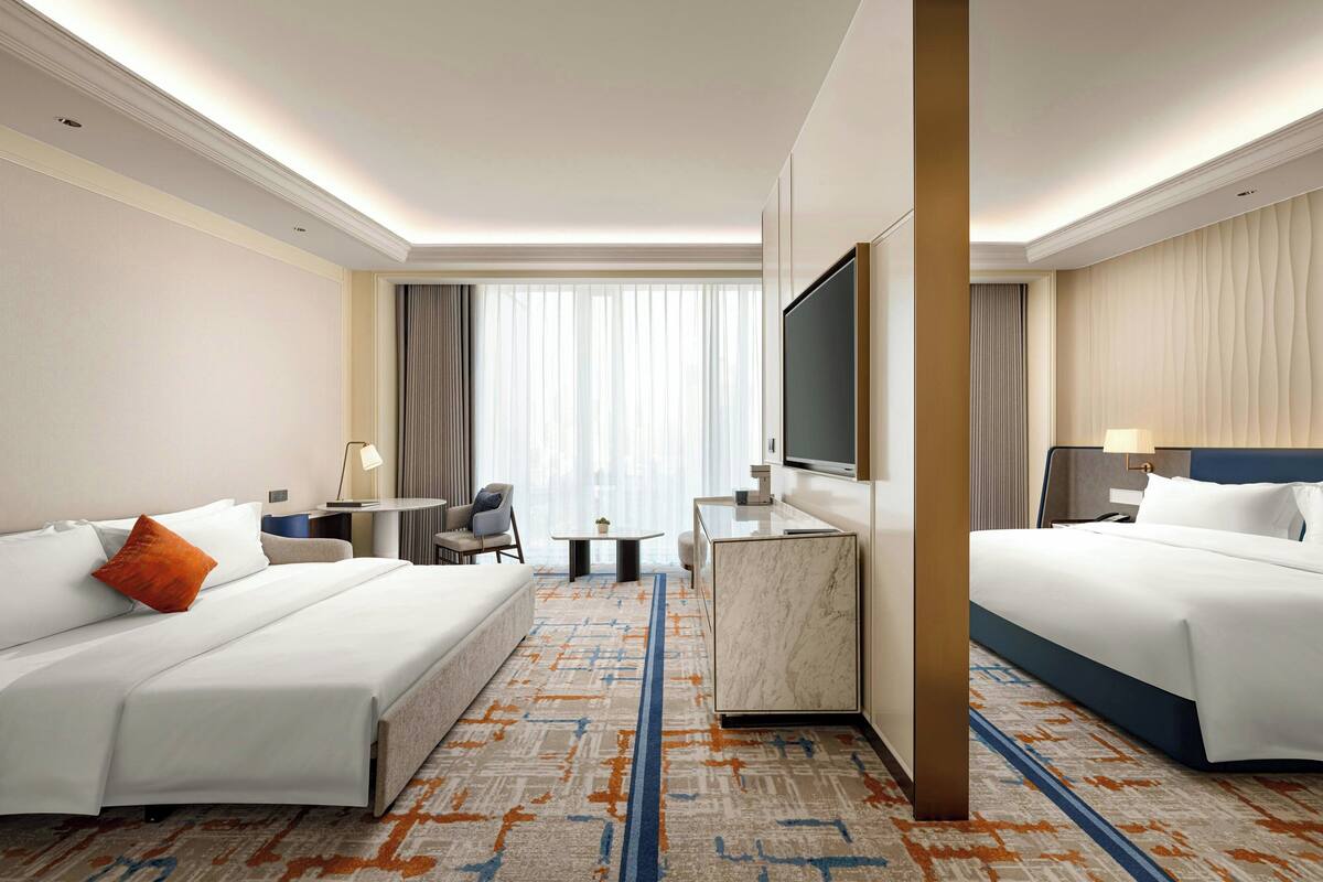 A hotel room with two beds, a desk, a TV, and a view of the city through large windows.