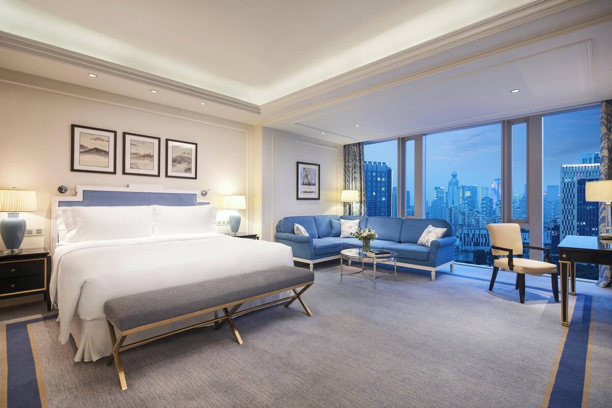 A modern hotel room with a large bed, a blue sofa, a small table, and a view of the city skyline through large windows.
