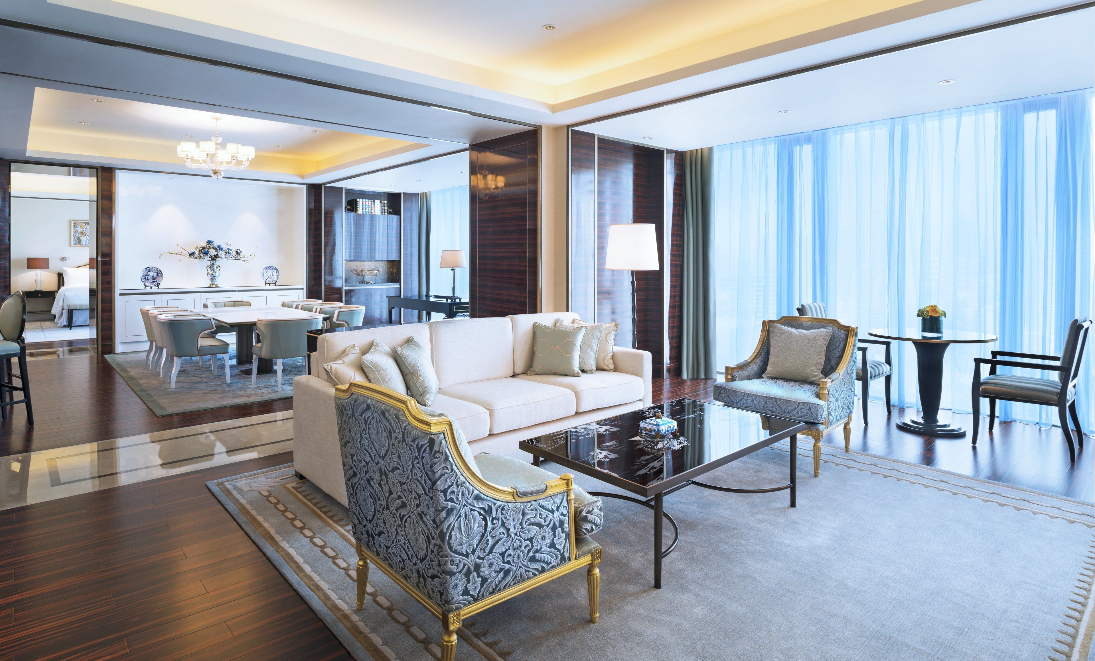 Un amplio vestíbulo de hotel con diseño moderno, que incluye un sofá blanco, sillones estampados, una mesa de centro y una zona de comedor con candelabro.