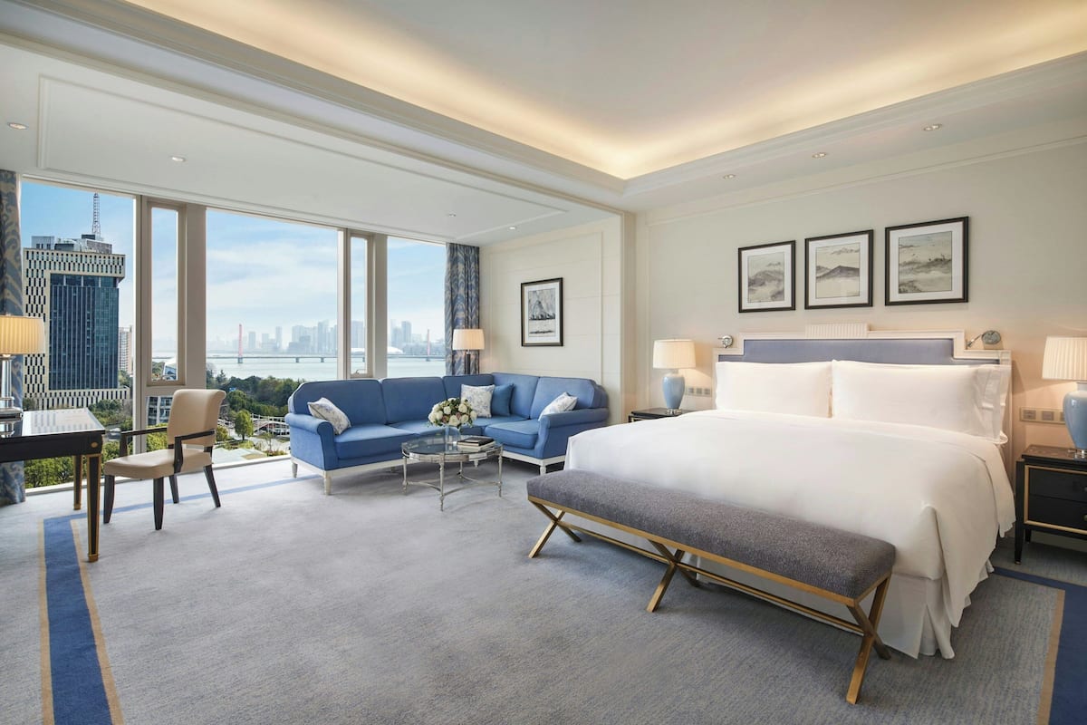 A modern hotel room with a large bed, a blue sofa, a small coffee table, and a view of the city skyline.