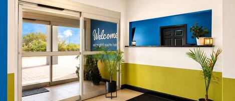 A hotel reception area with a glass door entrance, a welcome sign, potted plants, and a wall-mounted shelf with a plant and a small lamp.