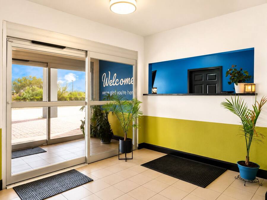 A hotel reception area with a glass door entrance, a welcome sign, potted plants, and a wall-mounted shelf with a plant and a small lamp.