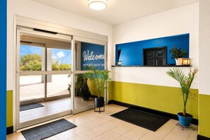 A hotel reception area with a glass door entrance, a welcome sign, potted plants, and a wall-mounted shelf with a plant and a small lamp. - Rodeway Inn Dilley I-35 (Dilley)