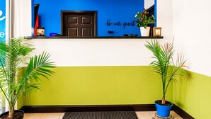 A reception area with a blue wall, a black door, and a 'be our guest' sign. There are two potted plants on either side of the reception desk.