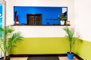 Une salle d’attente avec un mur bleu, une porte noire et une pancarte indiquant « Soyez les bienvenus ». Deux plantes en pot sont disposées de part et d’autre du comptoir d’accueil.