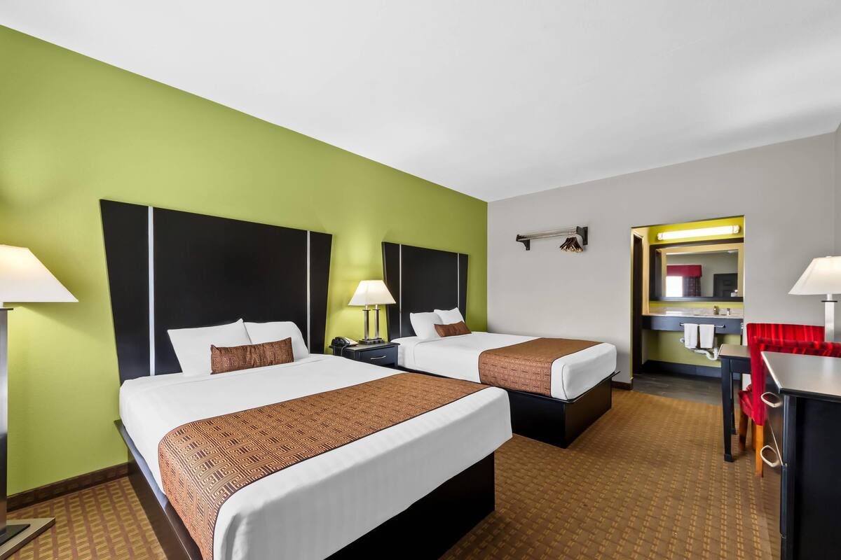 A hotel room with two beds, a desk, a chair, a mirror, and a bathroom with a sink and mirror.