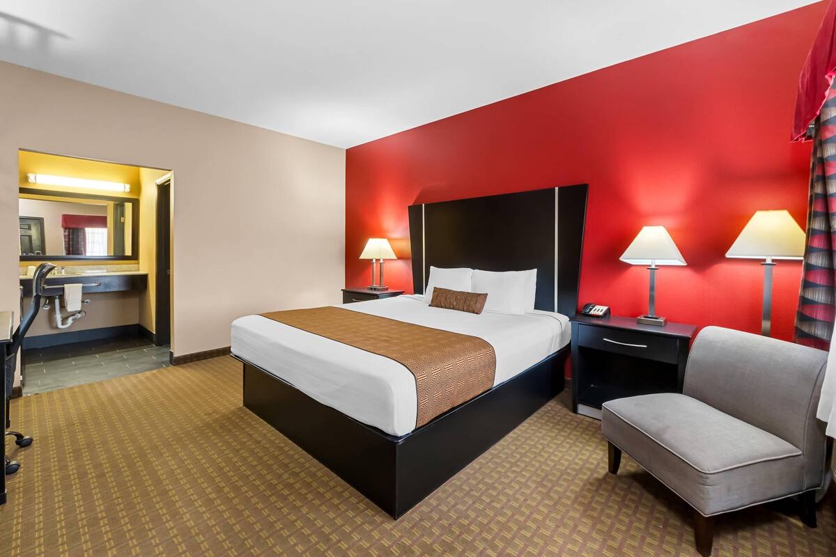 A hotel room with a bed, bedside tables, a chair, a desk, a mirror, and a red accent wall.