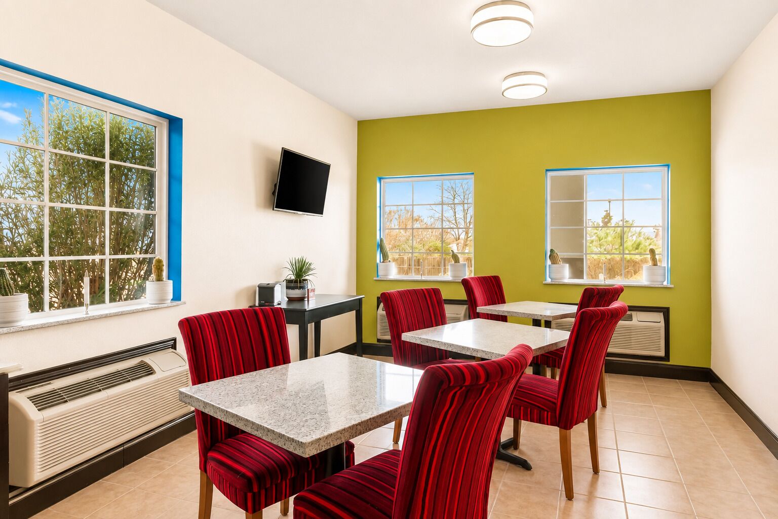 Un comedor con sillas tapizadas en rojo, una mesa con cubierta de mármol, un televisor de pantalla plana en la pared y un ventanal con vistas a árboles.