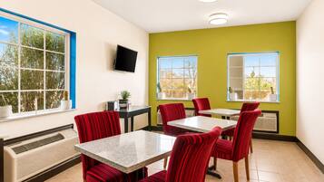 Un comedor con sillas tapizadas en rojo, una mesa con cubierta de mármol, un televisor de pantalla plana en la pared y un ventanal con vistas a árboles.