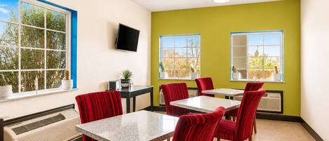 A dining area with red upholstered chairs, a marble-top table, a flat-screen TV mounted on the wall, and a window with a view of trees.