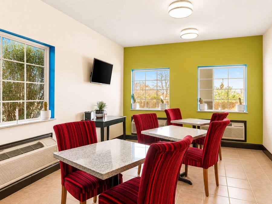 A dining area with red upholstered chairs, a marble-top table, a flat-screen TV mounted on the wall, and a window with a view of trees.