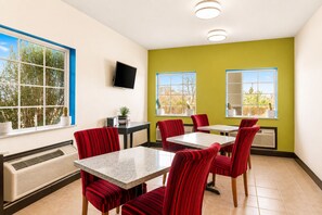 A dining area with red upholstered chairs, a marble-top table, a flat-screen TV mounted on the wall, and a window with a view of trees. - Rodeway Inn Dilley I-35 (Dilley)
