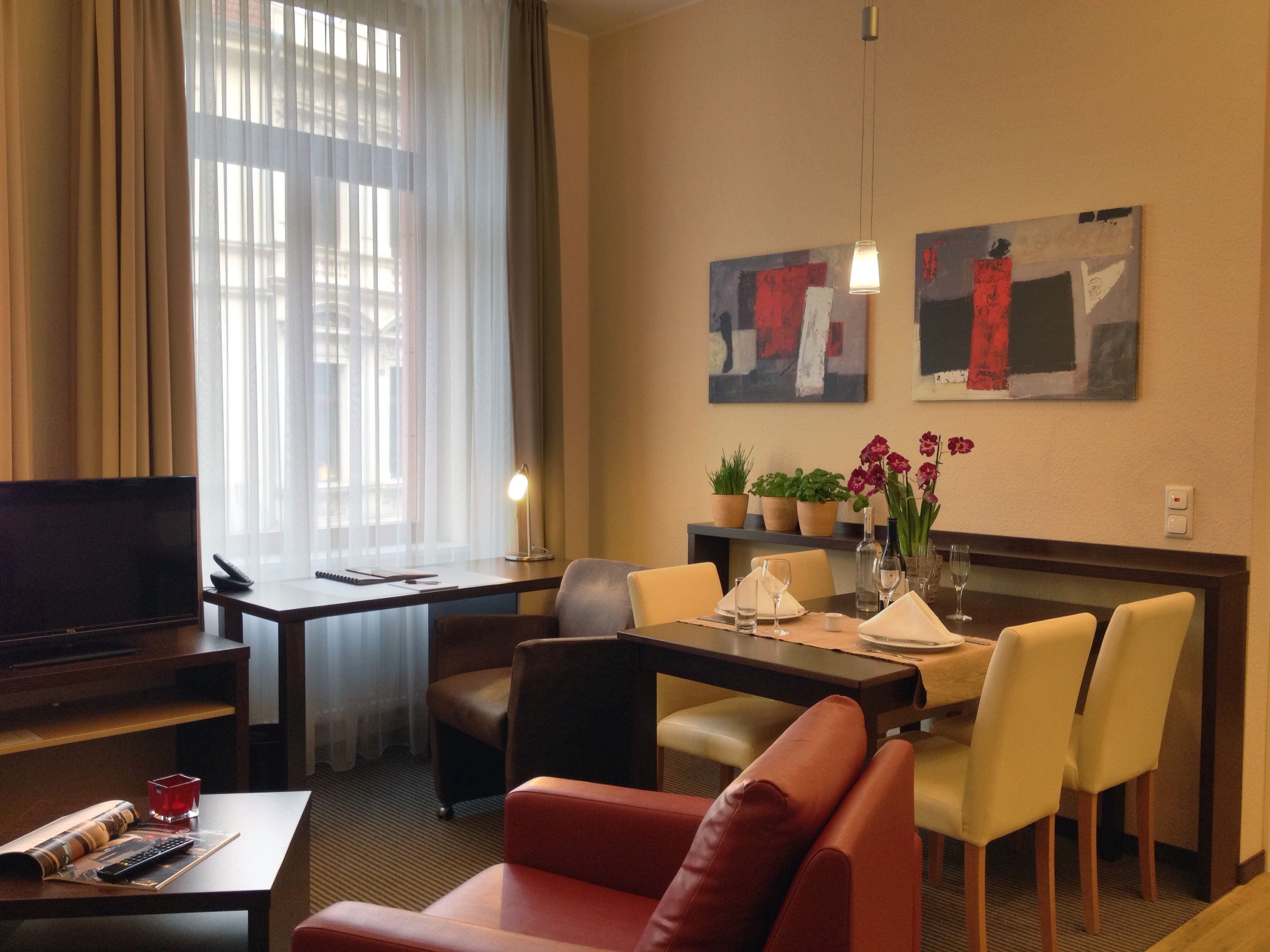 A hotel room with a TV, a dining table with chairs, a sofa, and artwork on the wall.