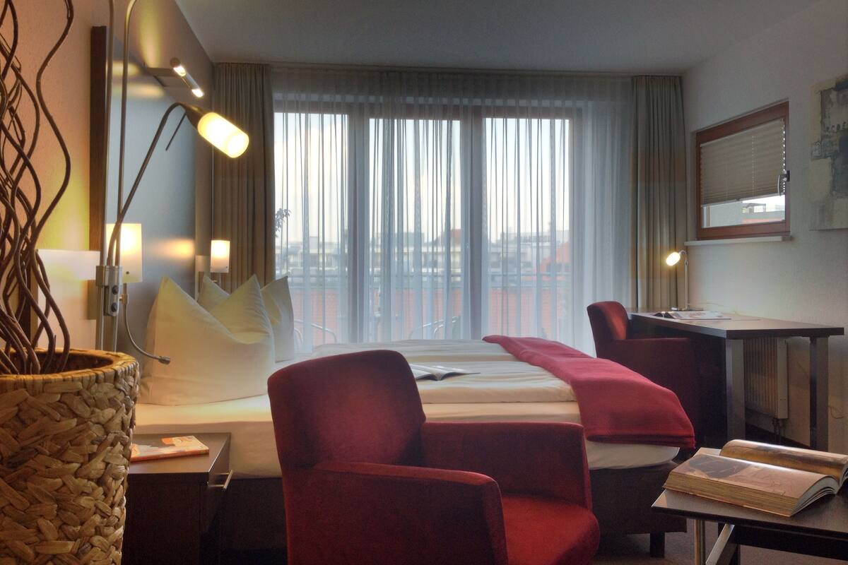 Habitación de hotel con cama, dos sillones rojos, un escritorio con lámpara, una ventana grande con cortinas y vistas a la ciudad.