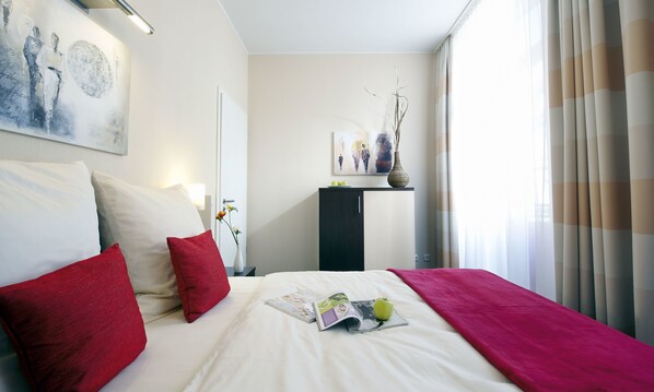 A neatly made bed with red and white pillows, a magazine, and an apple on the bed. - Apartmenthotel Quartier M (Leipzig)