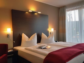 Una habitación de hotel con una cama grande, dos lámparas de noche, una mesita de noche, una silla roja y una ventana con cortinas.