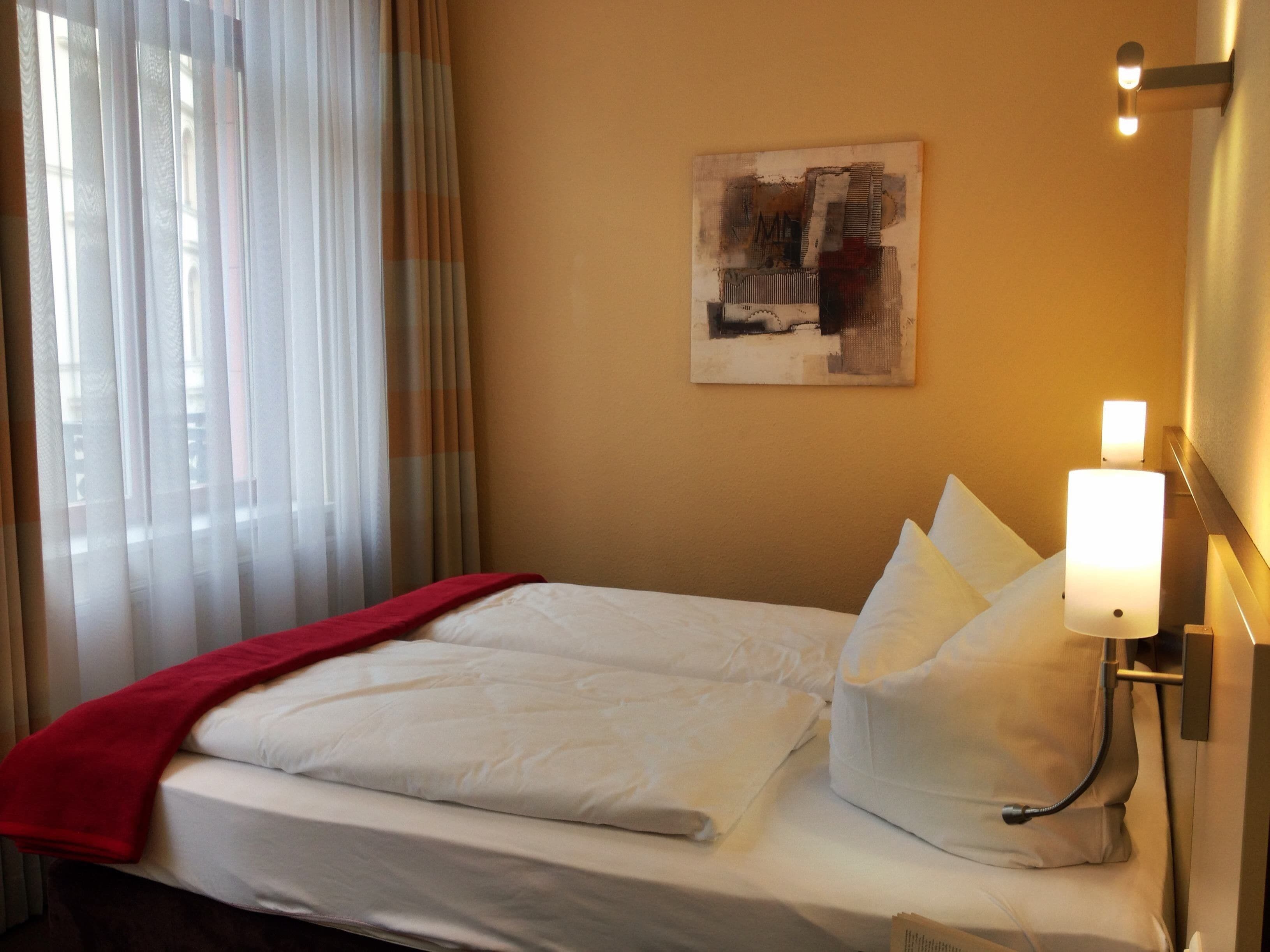 A hotel room with a bed, a red blanket, white pillows, a wall-mounted lamp, and a painting.