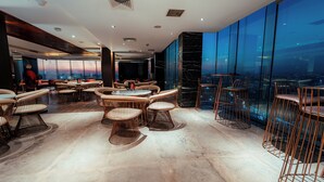A modern interior with round tables and wicker chairs, large windows offering a city view, and a marble floor. - The Peninsula Chittagong (Chittagong)