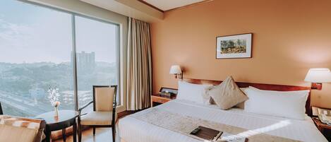 Habitación de hotel con un gran ventanal, una cama con sábanas blancas, una mesita de noche con una lámpara y una silla.