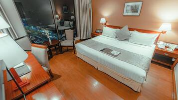 A hotel room with a large bed, a desk, a chair, and a view of the city at night.