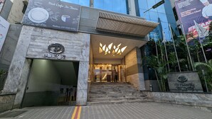 A modern building entrance with a staircase leading up to a glass door. There is a sign with a phone number and a logo on the wall. - The Peninsula Chittagong (Chittagong)