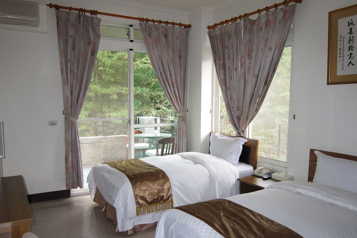 A hotel room with two beds, a balcony, and a view of greenery.