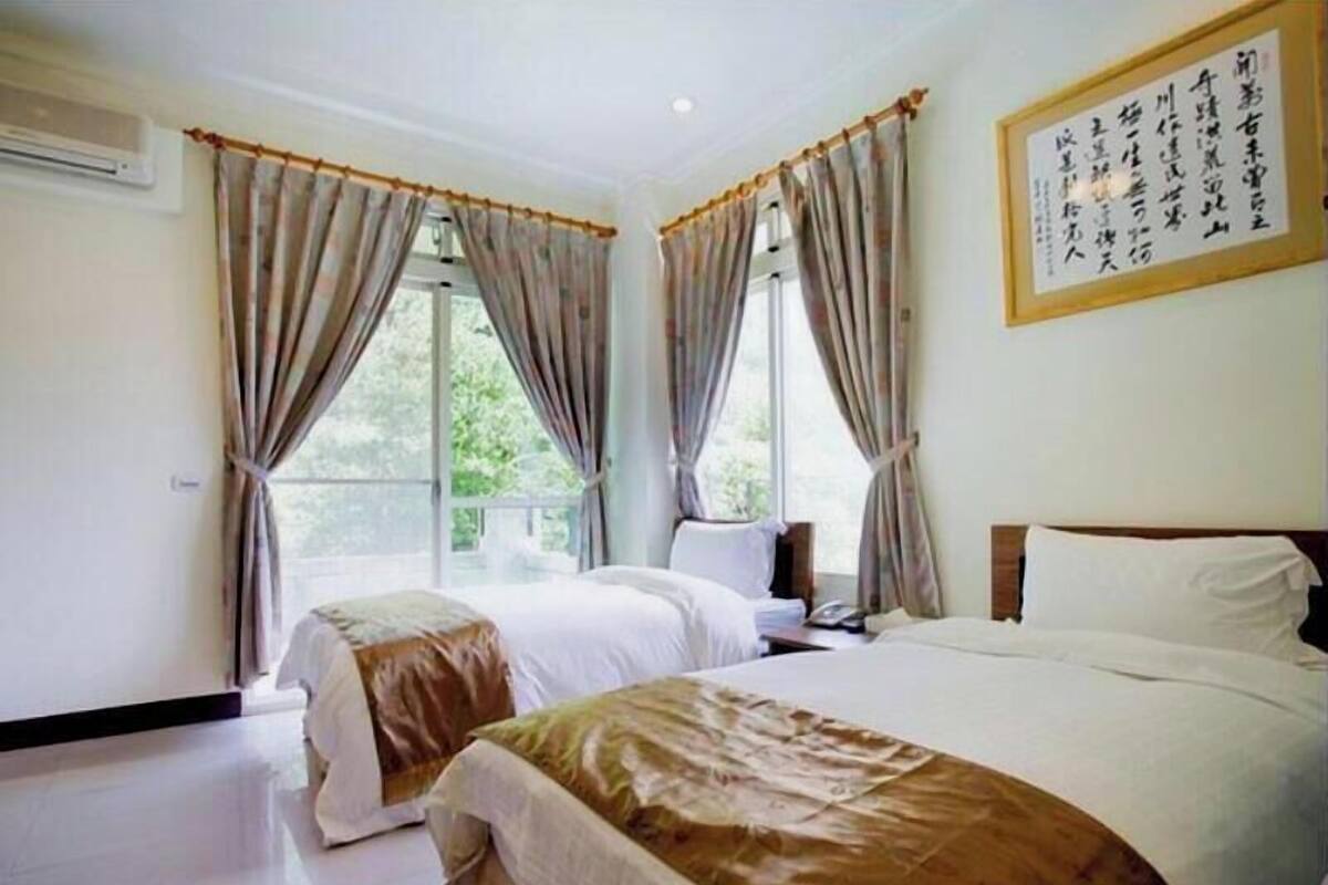A hotel room with two beds, a large window with curtains, an air conditioning unit, and a framed calligraphy piece on the wall.
