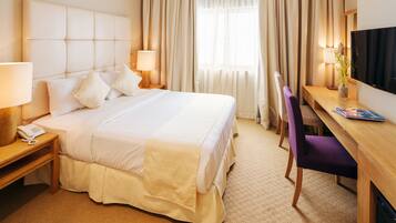 A hotel room with a large bed, bedside tables with lamps, a desk with a chair, a television, and a window with curtains.