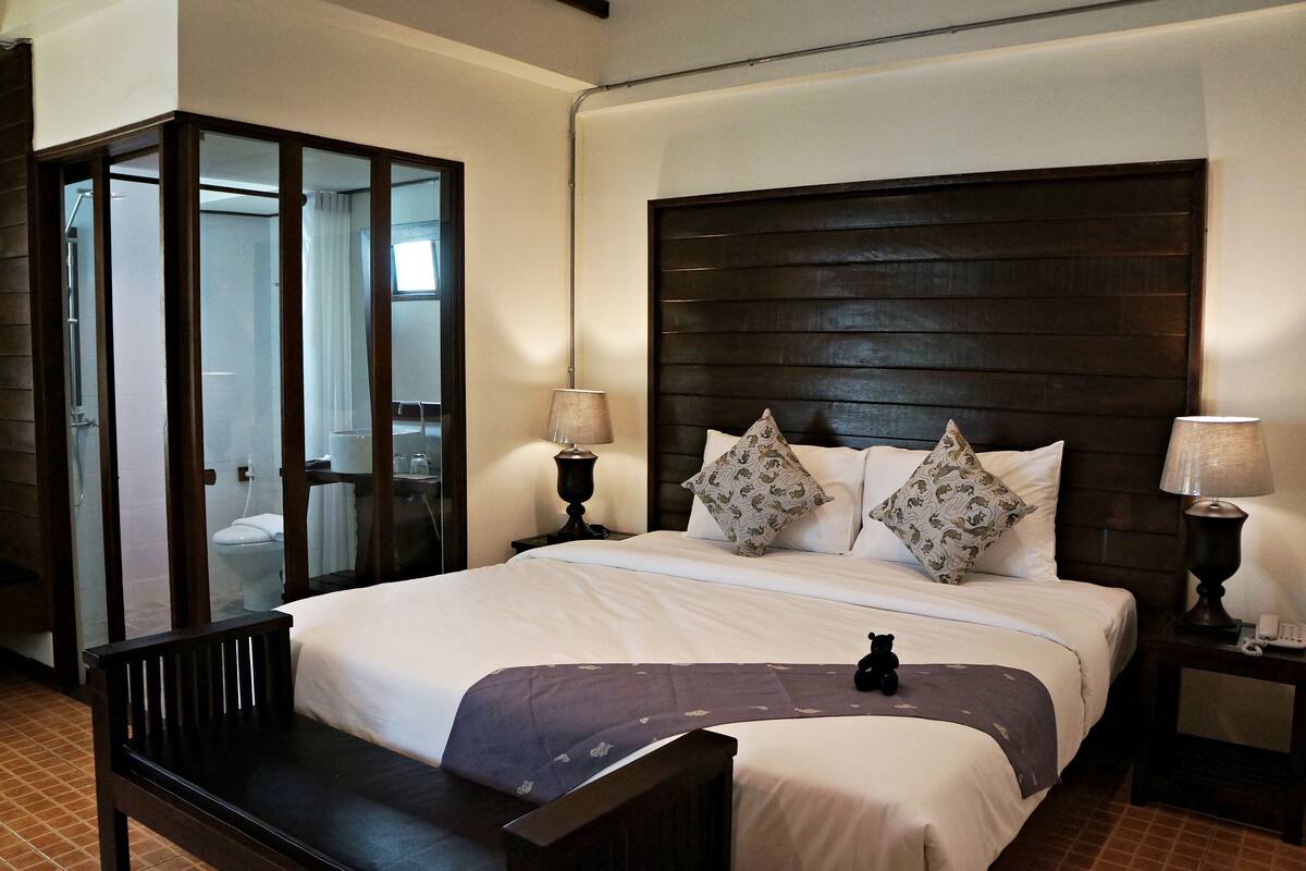 Habitación de hotel con una cama grande, dos lámparas de noche, un cabecero de madera y una zona de ducha con mampara de cristal.
