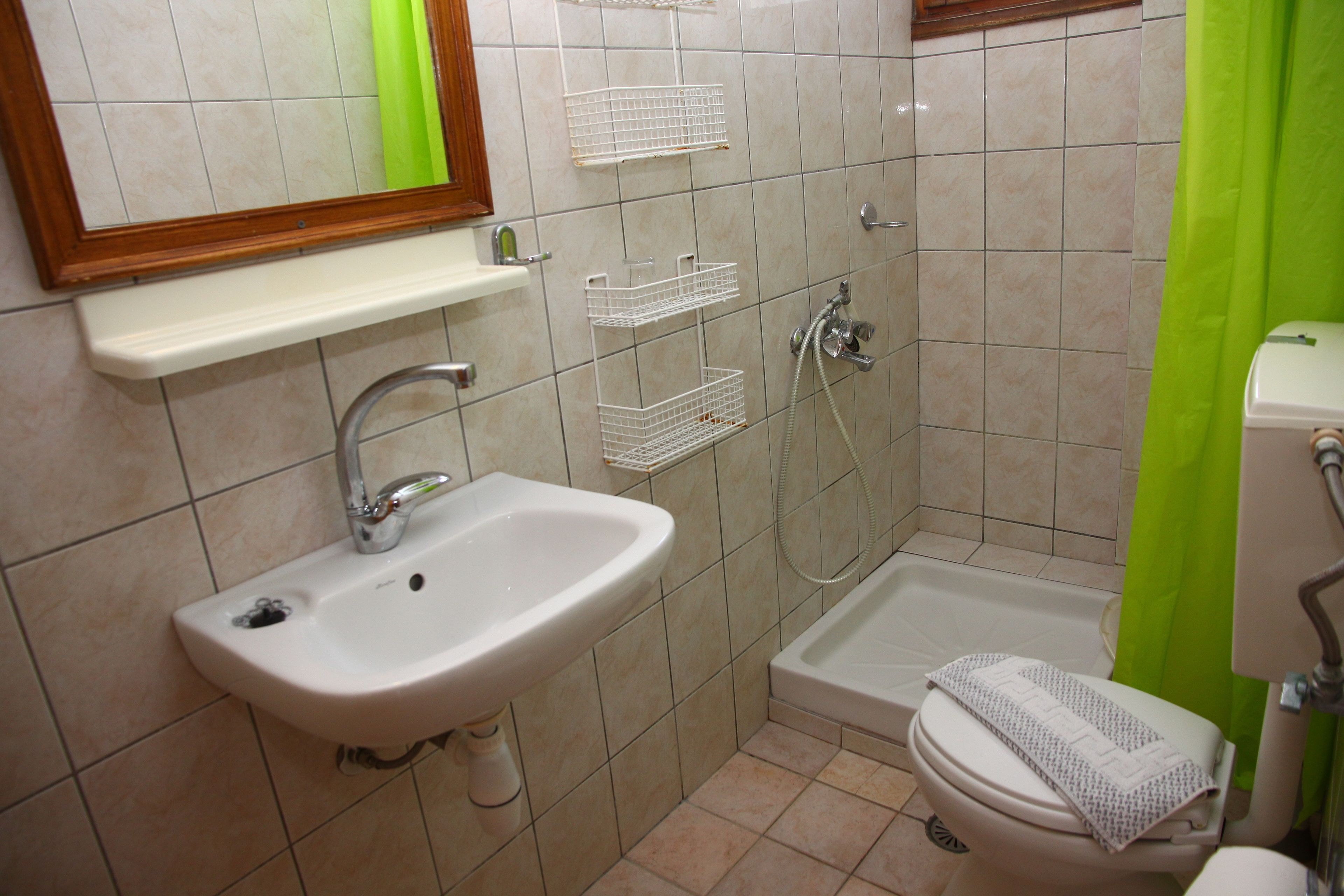 A bathroom with a sink, toilet, and shower. There are white shelves above the sink and a green shower curtain.