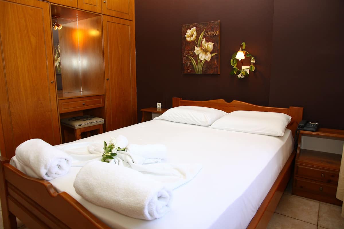 A neatly made bed with white linens and towels, a wooden wardrobe, a nightstand with a lamp, and a painting on the wall.