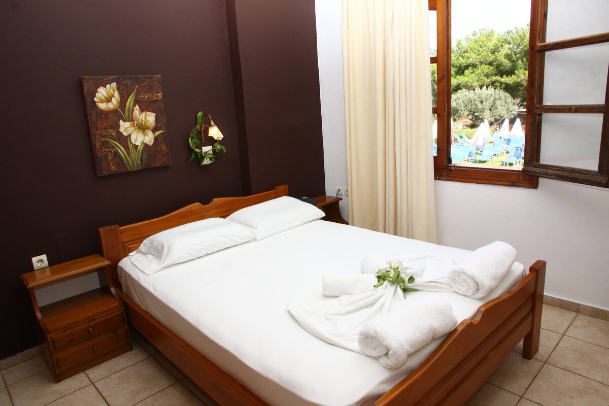 A neatly made bed with white linens and towels, a wooden nightstand with a lamp, and a painting of flowers on the wall.
