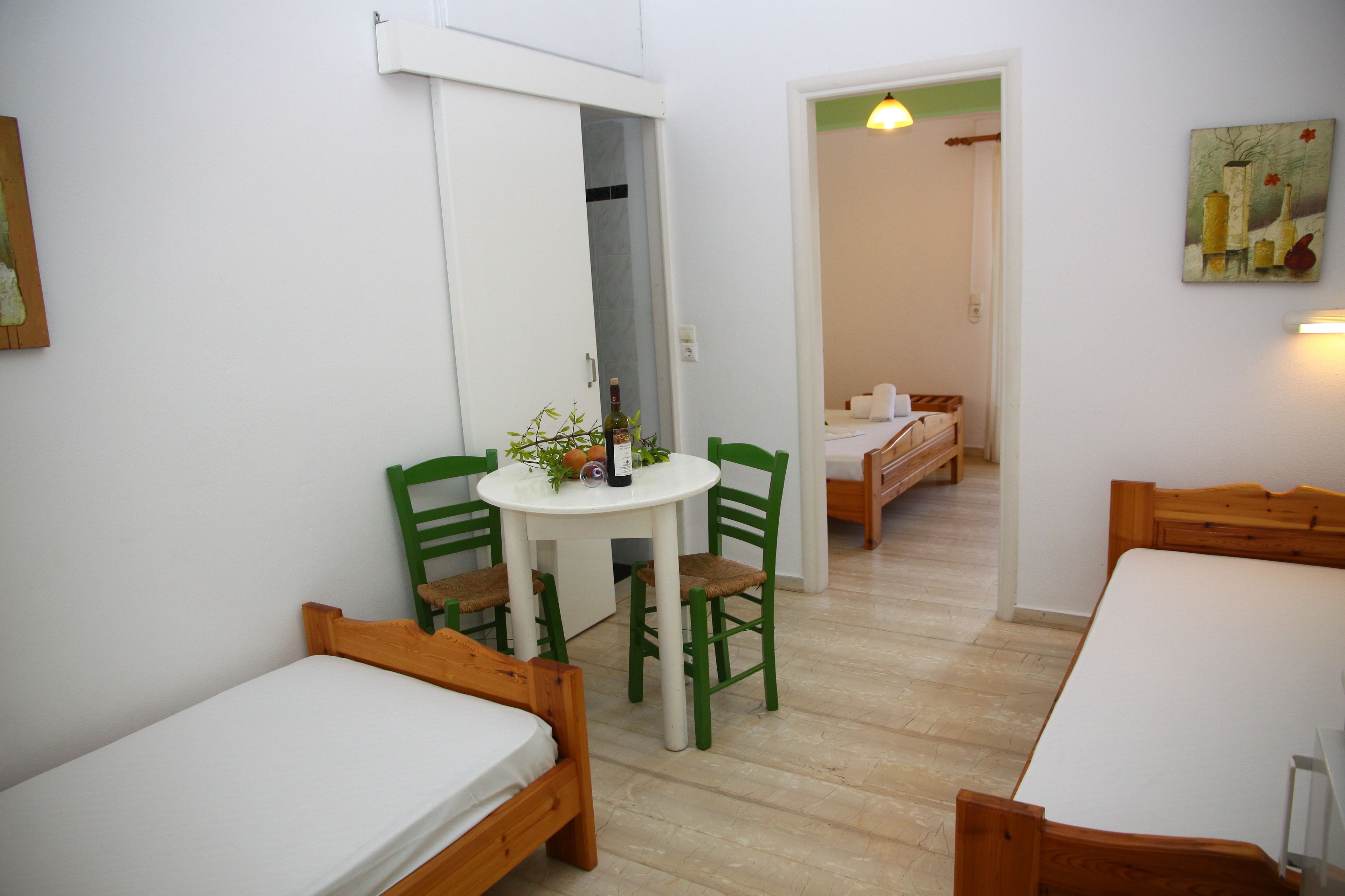 Une petite pièce propre, avec un lit, une table et des chaises, et une porte qui donne sur une autre pièce.
