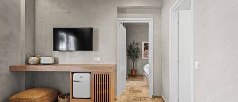 A hallway with a wooden bench, a small refrigerator, a television mounted on the wall, and a door leading to another room.