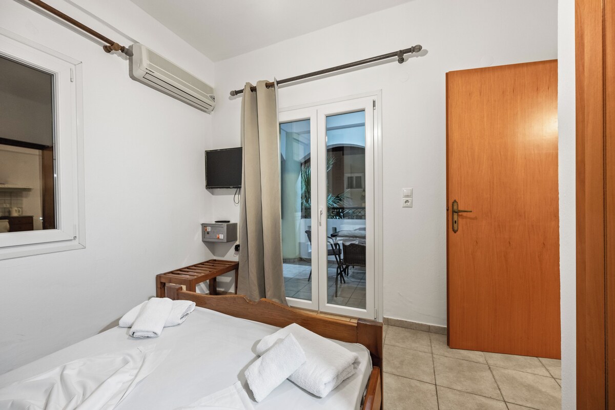 A hotel room with a bed, a TV, a desk, and a sliding glass door to a balcony.