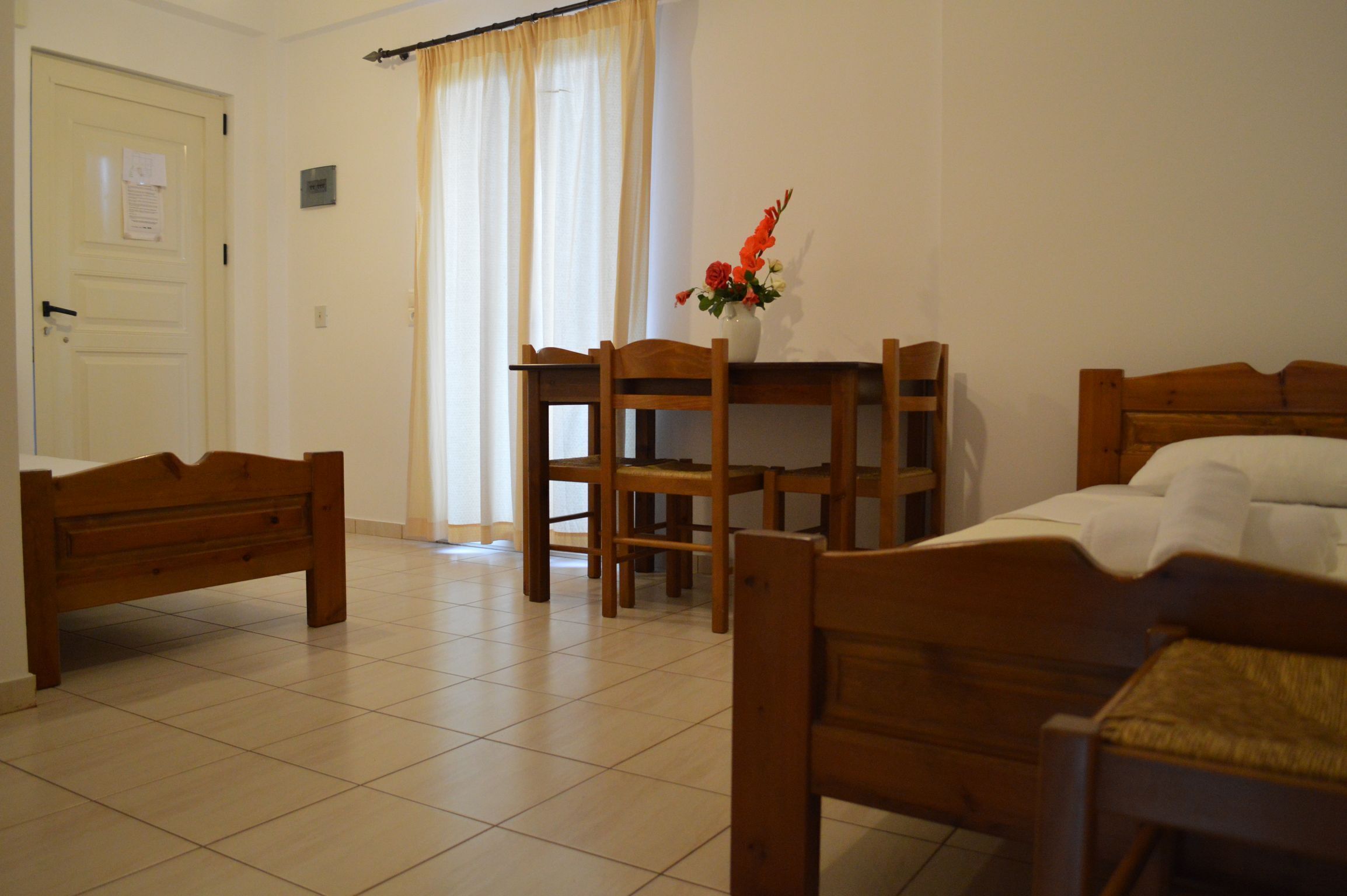 A room with a bed, a wooden chair, a small table with a vase of flowers, and a door with a notice on it.