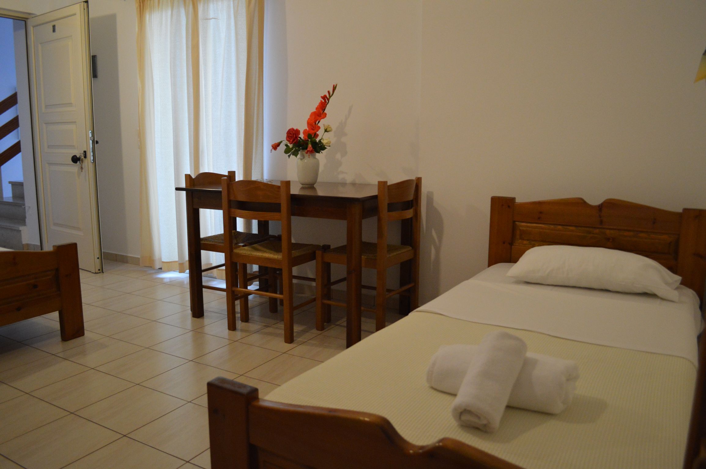 A hotel room with a bed, a wooden table with chairs, a vase with flowers, and a window with curtains.