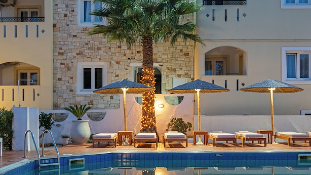 A poolside area with lounge chairs and umbrellas, a palm tree, and a stone building facade.