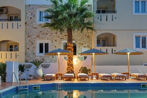 A poolside area with lounge chairs and umbrellas, a palm tree, and a stone building facade. - Nikos Apartments (Hersonissos)
