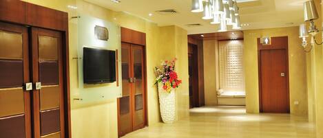 A hotel lobby with wooden doors, a flat-screen TV, a white vase with flowers, and a chandelier.