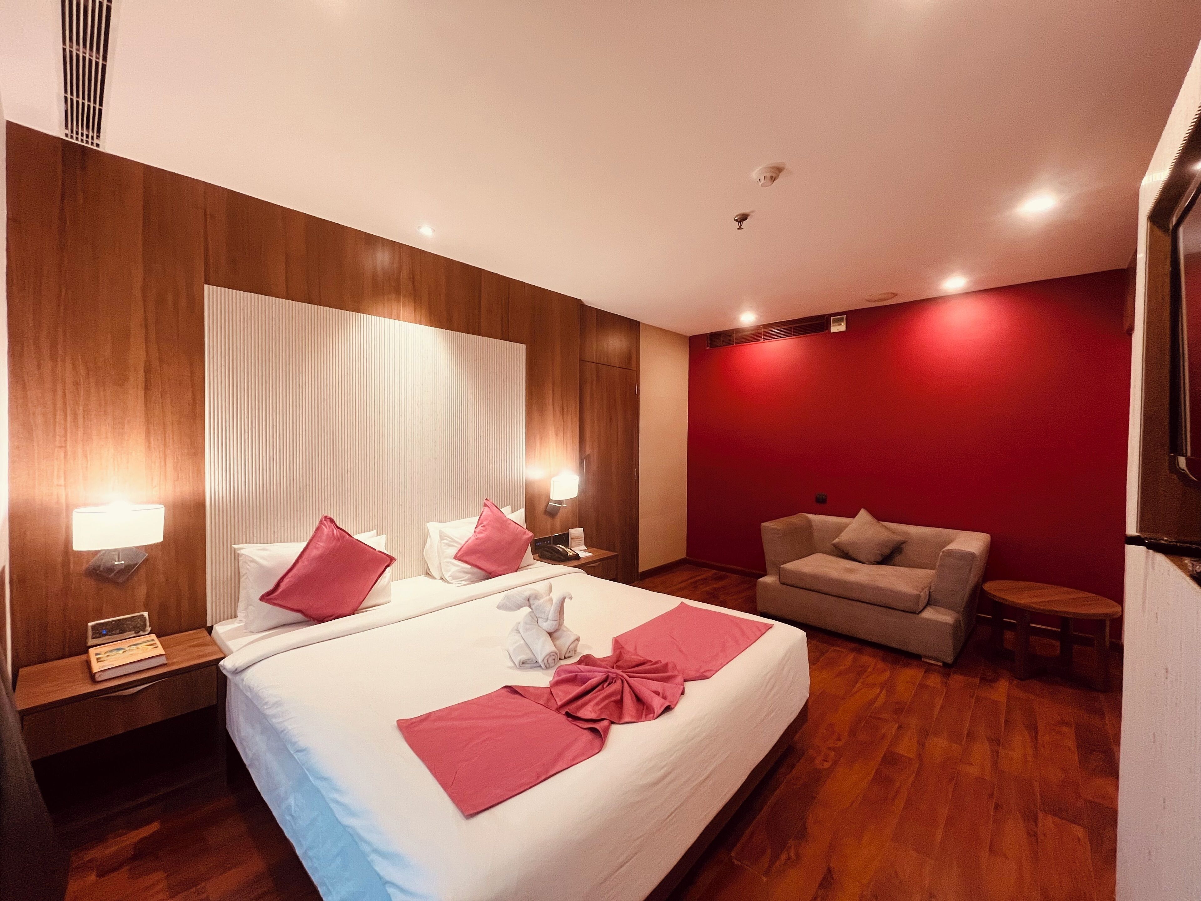 Habitación de hotel con una cama grande, una pared de acento roja, piso de madera, un pequeño sofá y una mesita de noche con una lámpara y un libro.