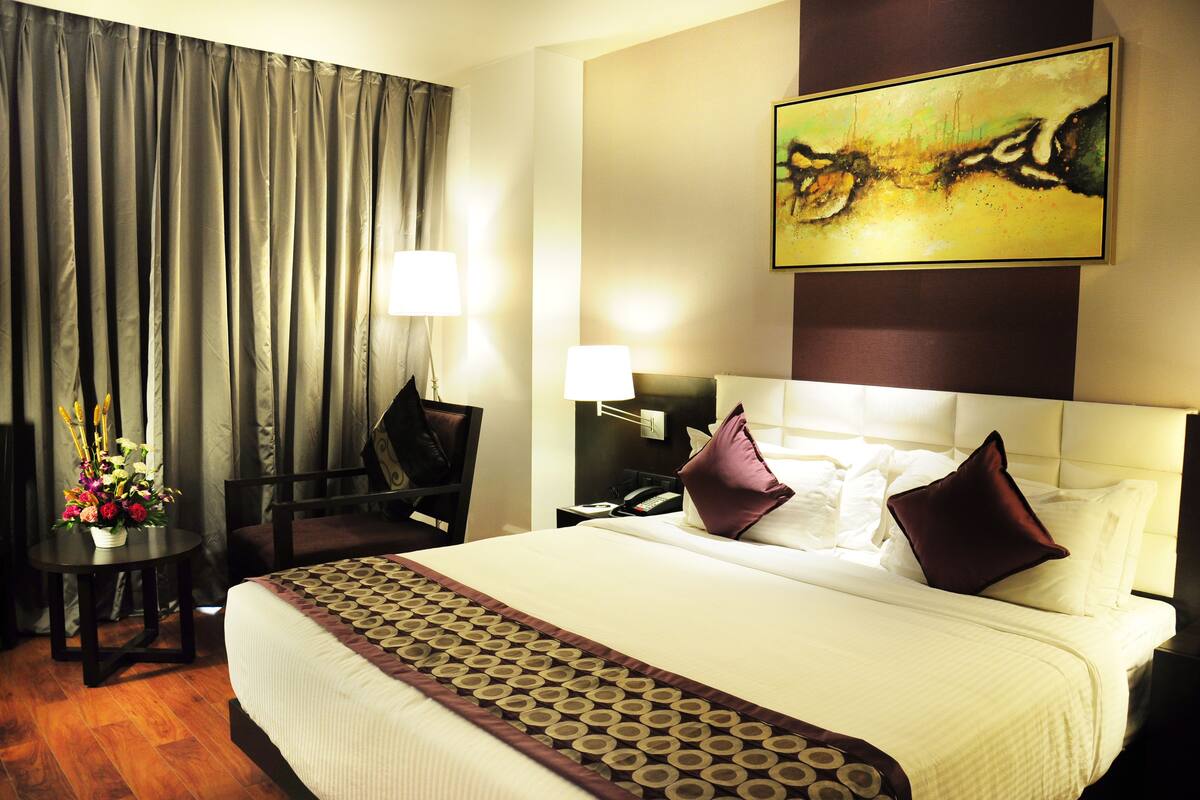 A hotel room with a large bed, a desk with a lamp, a chair, and a painting on the wall.