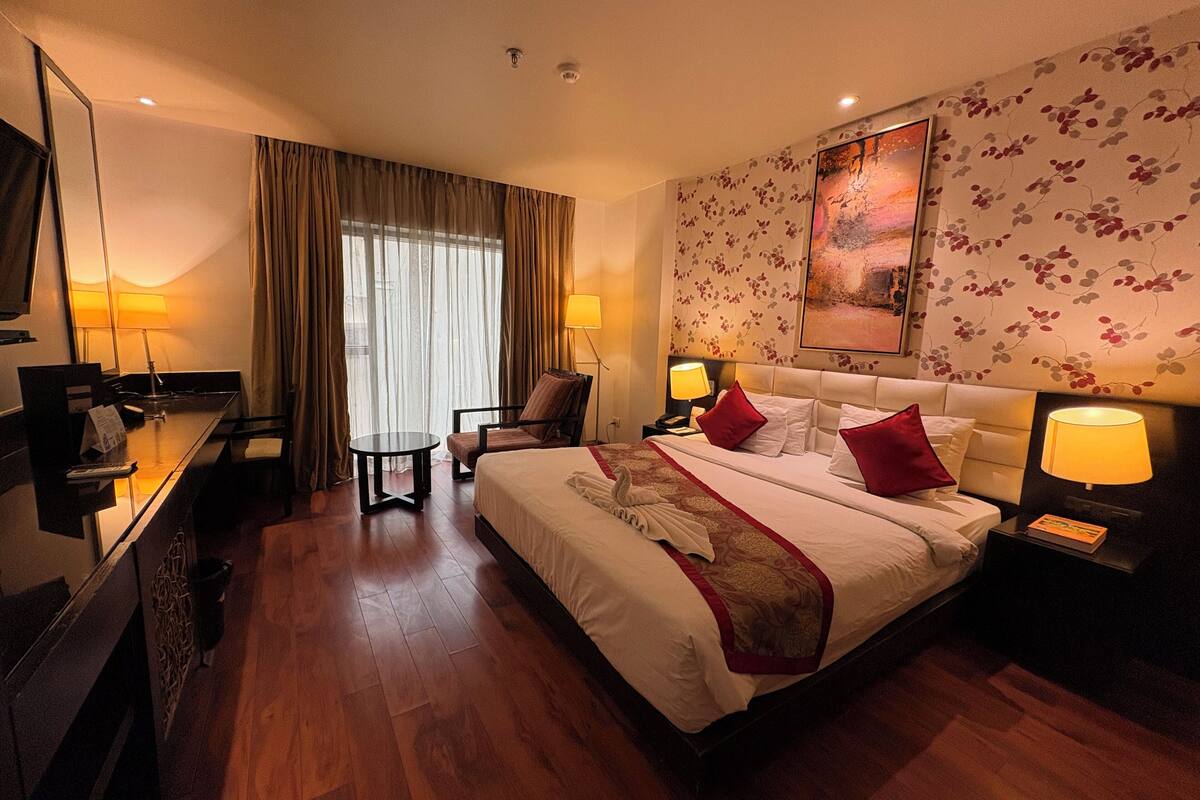 A hotel room with a large bed, a desk, a chair, a small table, and a large window with curtains.