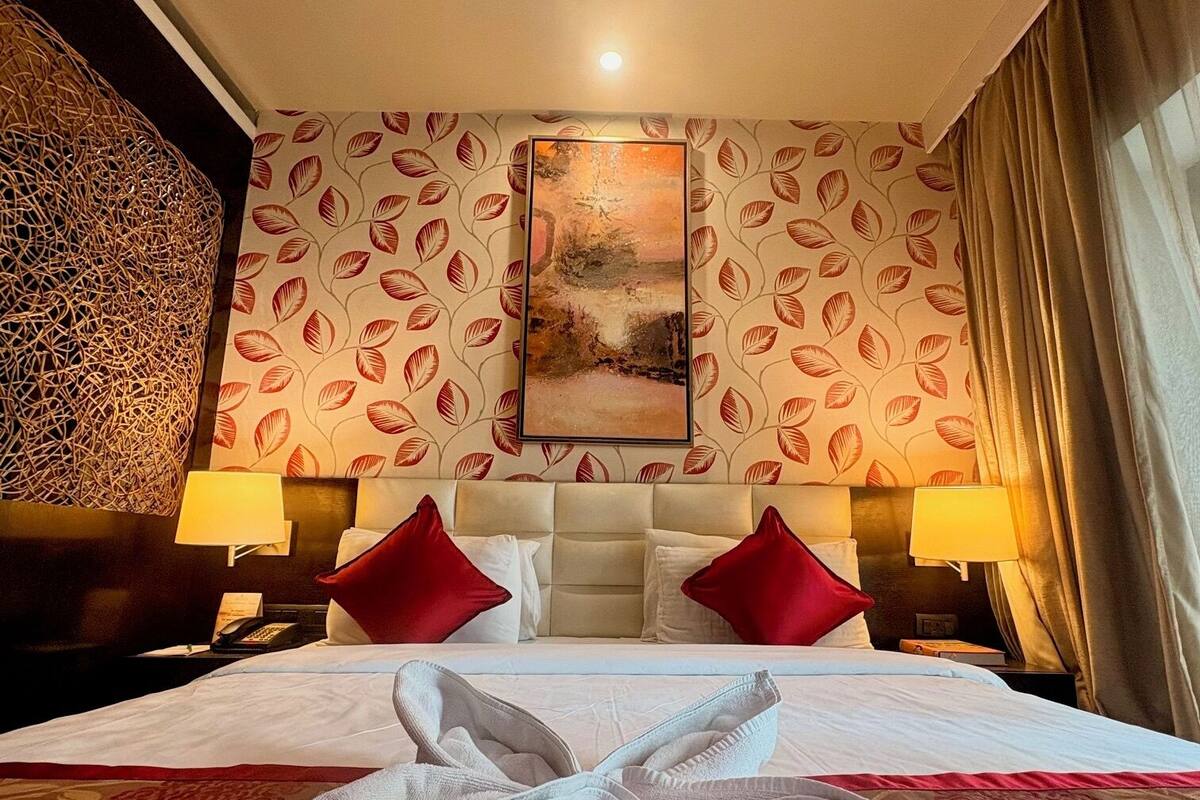 A hotel room with a large bed, two red pillows, a headboard, a painting on the wall, and a decorative wall panel.
