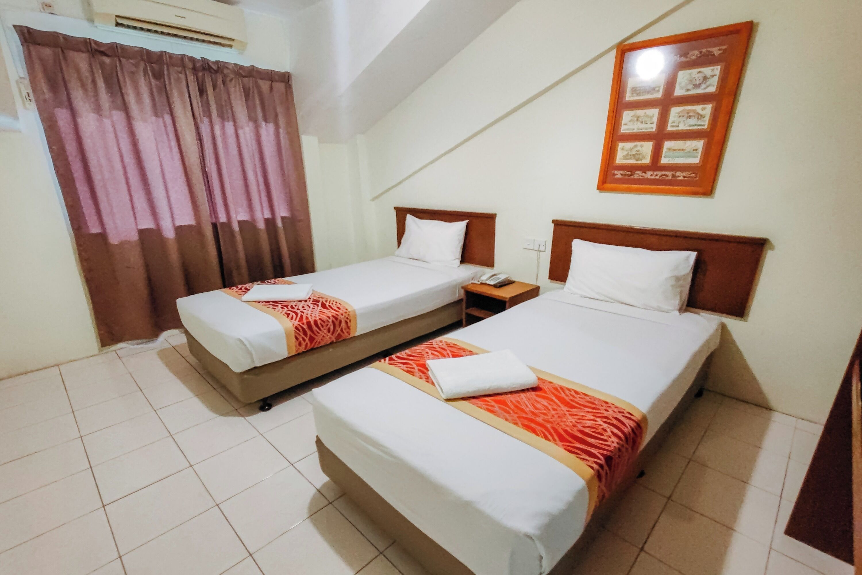 A hotel room with two single beds, a small bedside table with a lamp, a wall-mounted air conditioner, and a framed picture on the wall.