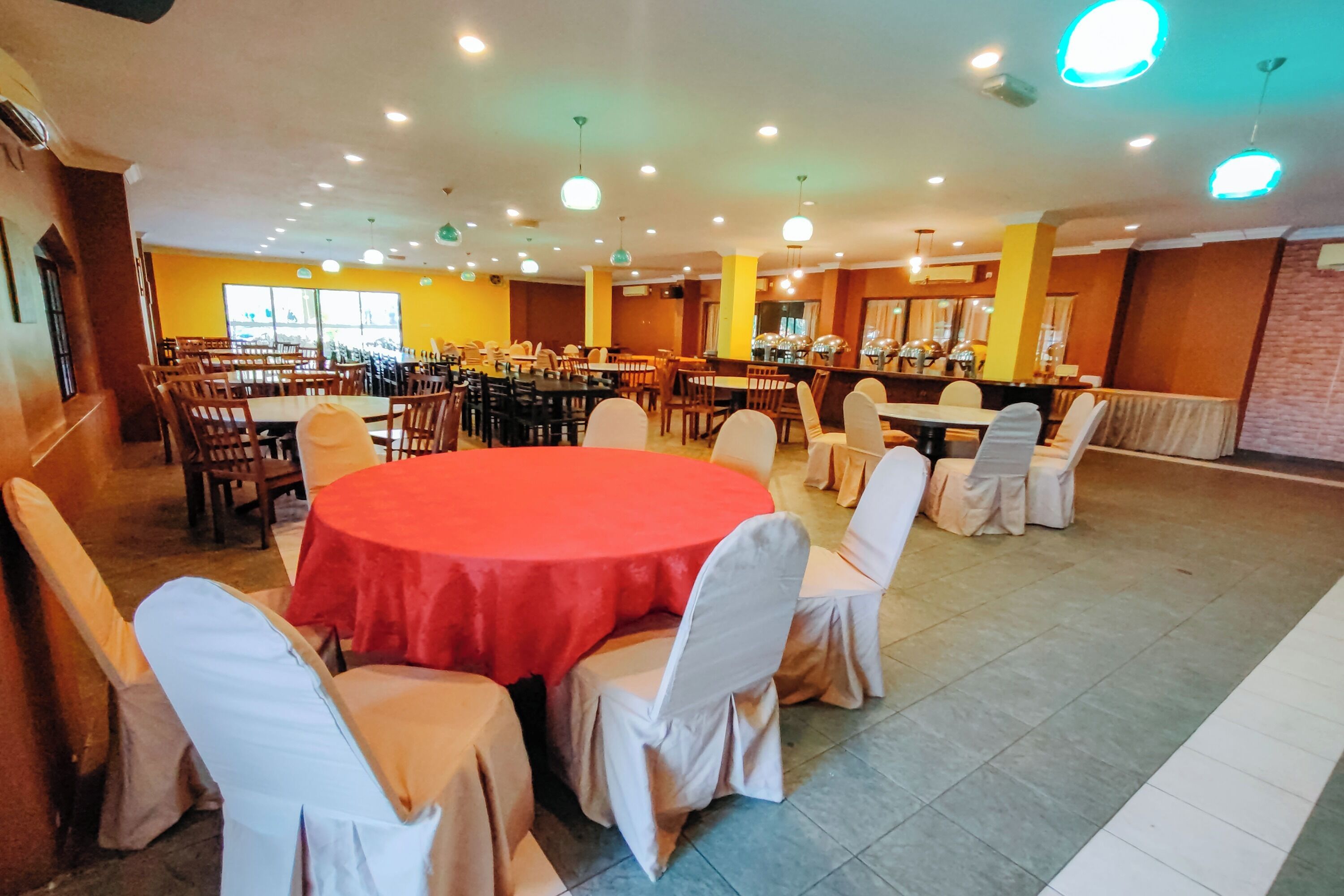 A spacious dining area with multiple tables and chairs, a central round table with a red tablecloth, and a bright, well-lit environment.