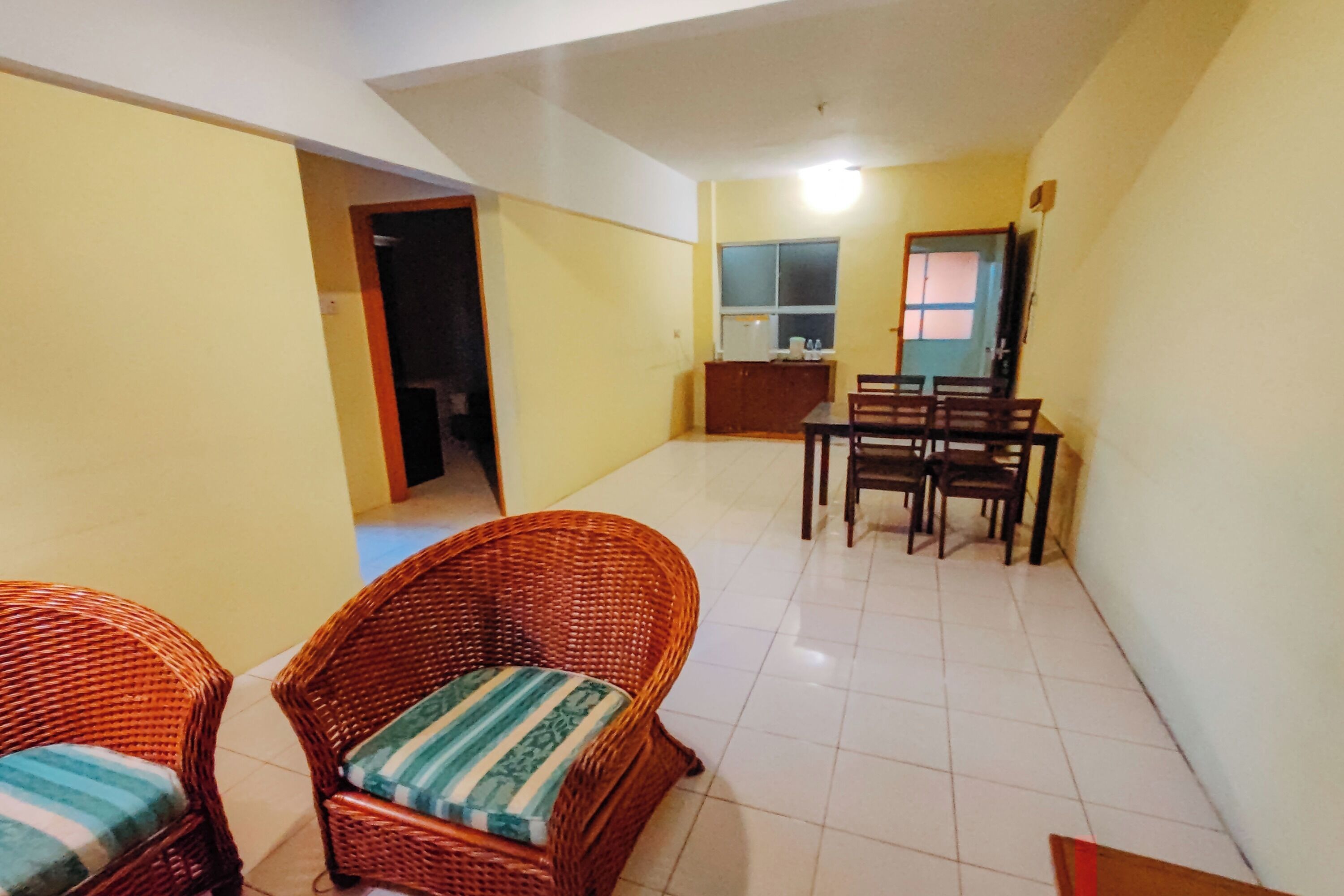 A room with two wicker chairs, a dining table with chairs, a small kitchenette, and a door leading to another room.